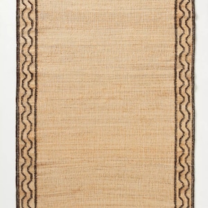 May include: A natural fiber rug with a brown and black geometric border. The rug has a woven texture and a light brown color.