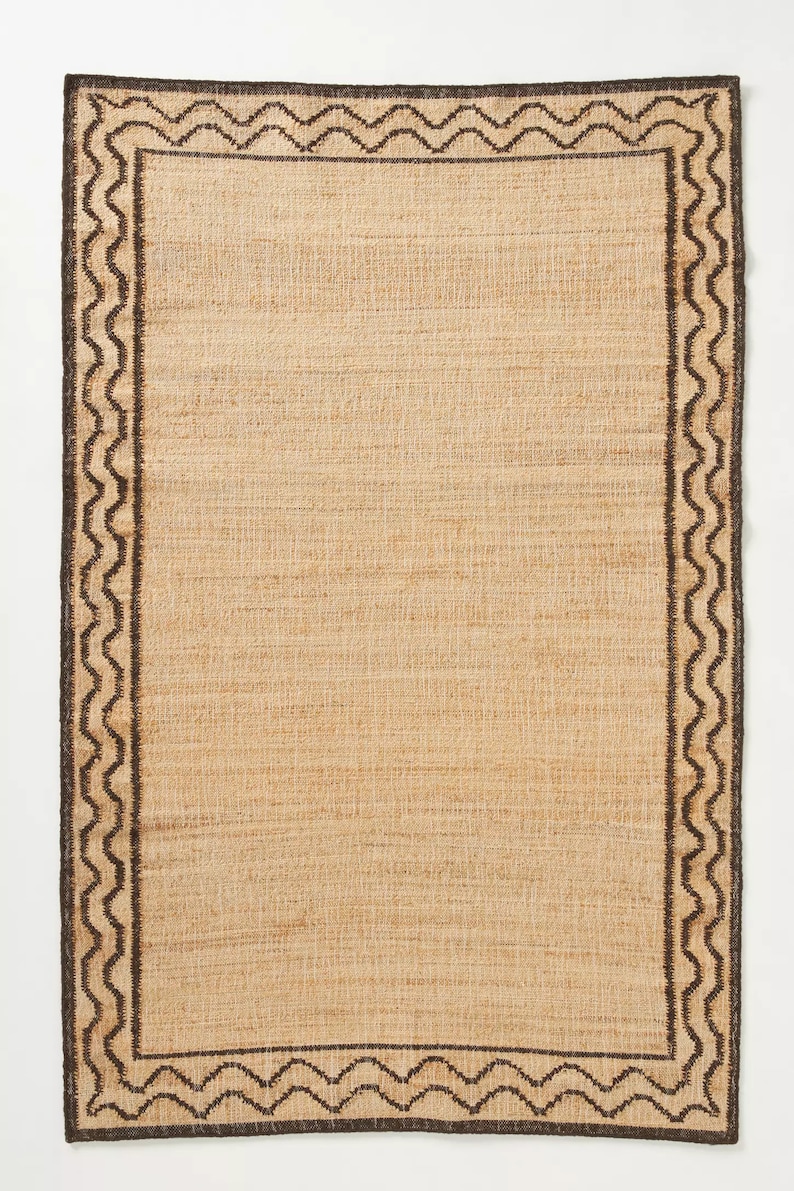 May include: A natural fiber rug with a brown and black geometric border. The rug has a woven texture and a light brown color.