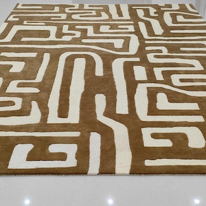 May include: A beige and brown area rug with a geometric pattern of interlocking lines and shapes.