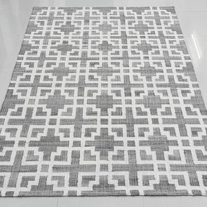 May include: A rectangular gray and white patterned rug. The rug features a geometric design with repeating cross-like shapes. The rug is made of a woven material and is placed on a white tile floor.