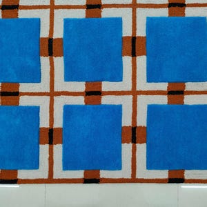 Op de afbeelding: Een geometrisch tapijt met een patroon van blauwe vierkanten in een raster van witte, bruine en zwarte lijnen. Het tapijt heeft een textuur en een modern design, geschikt voor interieurdecoratie.