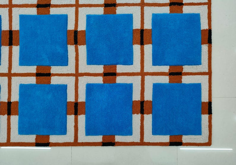 Op de afbeelding: Een geometrisch tapijt met een patroon van blauwe vierkanten in een raster van witte, bruine en zwarte lijnen. Het tapijt heeft een textuur en een modern design, geschikt voor interieurdecoratie.