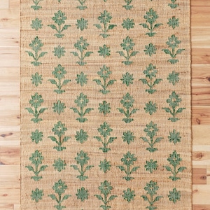 Op de afbeelding: Een vloerkleed van natuurlijke vezels met een groen bloemenpatroon. Het vloerkleed heeft een geweven textuur en een lichtbruine achtergrond.