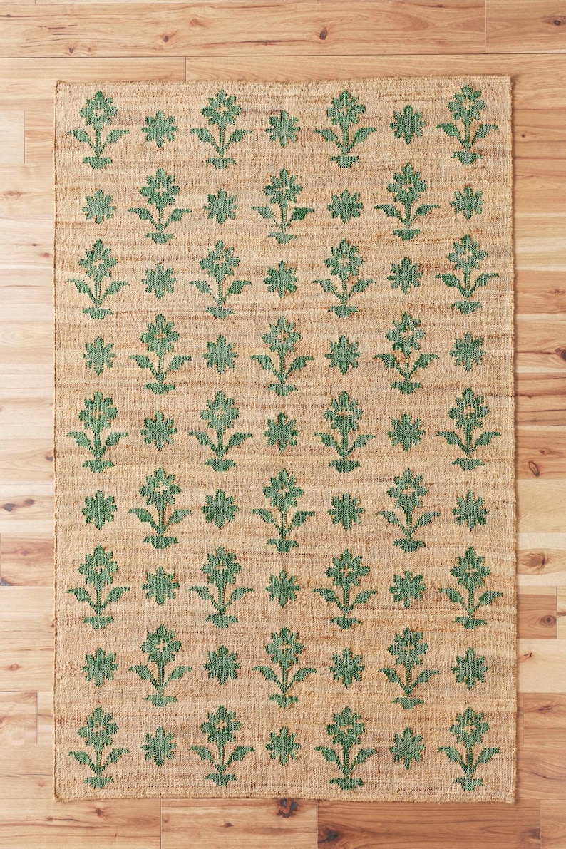 Op de afbeelding: Een vloerkleed van natuurlijke vezels met een groen bloemenpatroon. Het vloerkleed heeft een geweven textuur en een lichtbruine achtergrond.