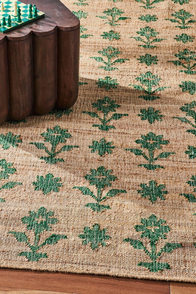 Op de afbeelding: Een tan en groen vloerkleed met een bloemenpatroon. Het kleed is gemaakt van een natuurlijke vezel en heeft een geweven textuur.