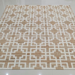 May include: A rectangular rug with a geometric pattern. The rug features a repeating design of white squares and rectangles against a tan background. The rug is made of a woven material and is placed on a light-colored floor.