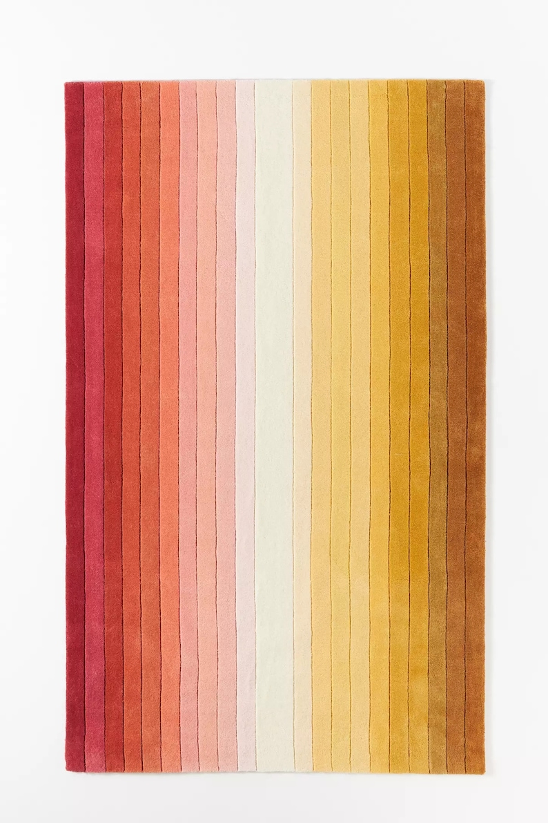 K&ouml;nnte beinhalten: Ein bunter gestreifter Teppich in Rot-, Orange-, Rosa-, Gelb- und Braunt&ouml;nen. Der Teppich ist aus weichem, pl&uuml;schigem Material gefertigt und hat ein modernes, geometrisches Design.