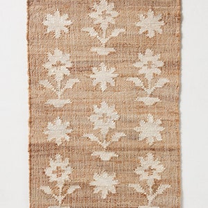 May include: A rectangular jute rug with a white floral pattern. The rug has a natural brown color and a woven texture.