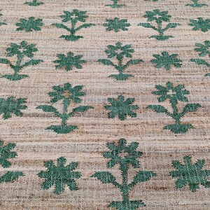 Op de afbeelding: Een close-up van een vloerkleed van natuurlijke vezels met een herhalend patroon van groene bloemenontwerpen. Het vloerkleed heeft een geweven textuur en een lichtbruine achtergrond.
