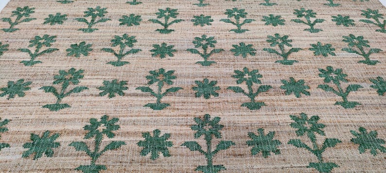 Op de afbeelding: Een close-up van een vloerkleed van natuurlijke vezels met een herhalend patroon van groene bloemenontwerpen. Het vloerkleed heeft een geweven textuur en een lichtbruine achtergrond.