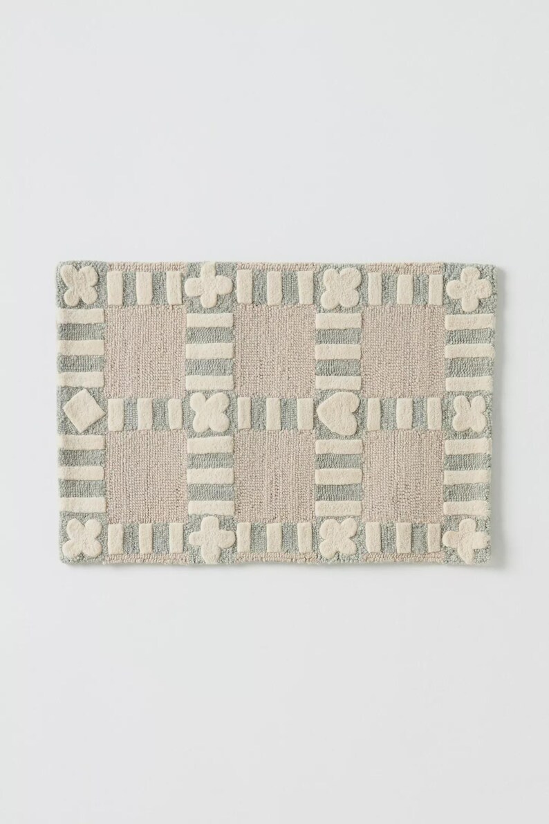 May include: Rectangular rug with a grid pattern. The rug features a light beige background with a border and grid lines in light gray. Decorative shapes like flowers and hearts are incorporated.
