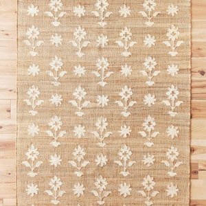 May include: A beige and white area rug with a floral pattern. The rug has a woven texture and features a repeating pattern of white flowers on a beige background.