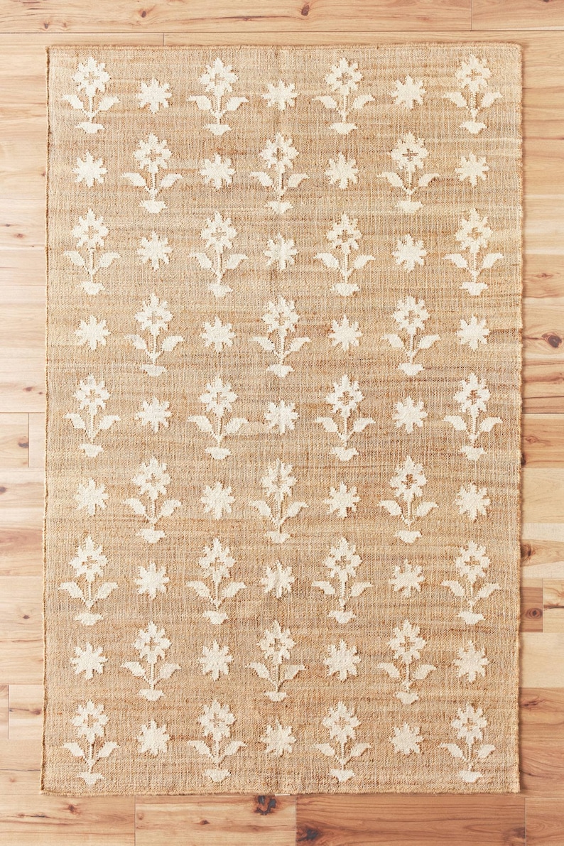 May include: A beige and white area rug with a floral pattern. The rug has a woven texture and features a repeating pattern of white flowers on a beige background.