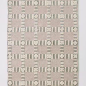 May include: A rectangular rug with a grid pattern. The rug features a light pink background with a repeating pattern of gray and off-white shapes, including hearts, diamonds, and four-leaf clovers. The rug is made of a soft, textured material.