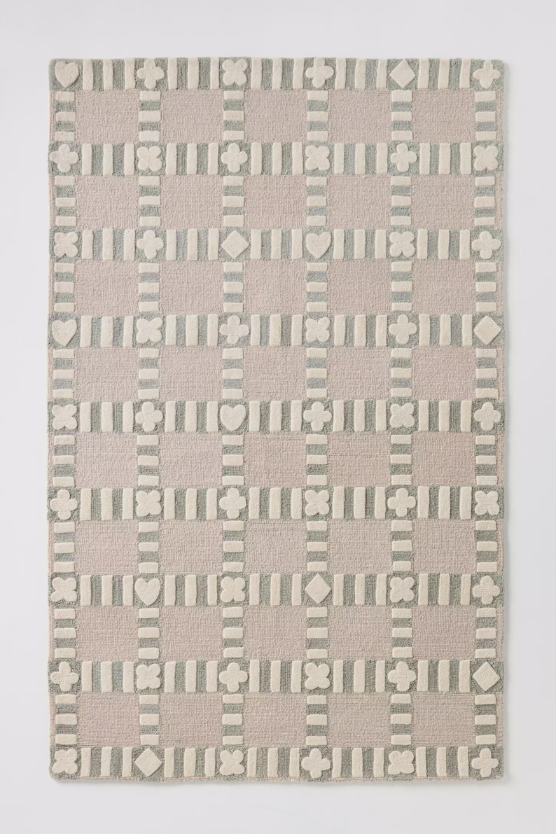 May include: A rectangular rug with a grid pattern. The rug features a light pink background with a repeating pattern of gray and off-white shapes, including hearts, diamonds, and four-leaf clovers. The rug is made of a soft, textured material.