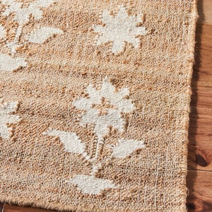 May include: Close-up of a natural fiber rug with a white floral pattern. The rug has a woven texture and a frayed edge.