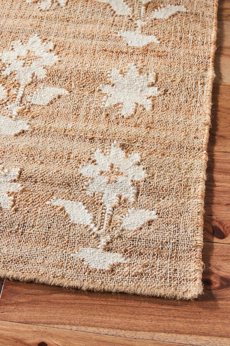 May include: Close-up of a natural fiber rug with a white floral pattern. The rug has a woven texture and a frayed edge.