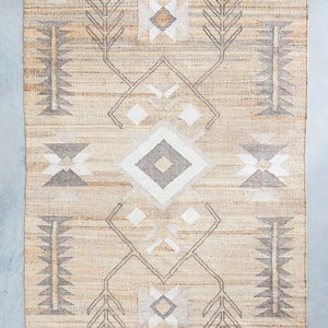 May include: A beige and gray area rug with a geometric pattern. The rug has a diamond shape in the center with smaller diamond shapes on either side. The rug has a fringe border.