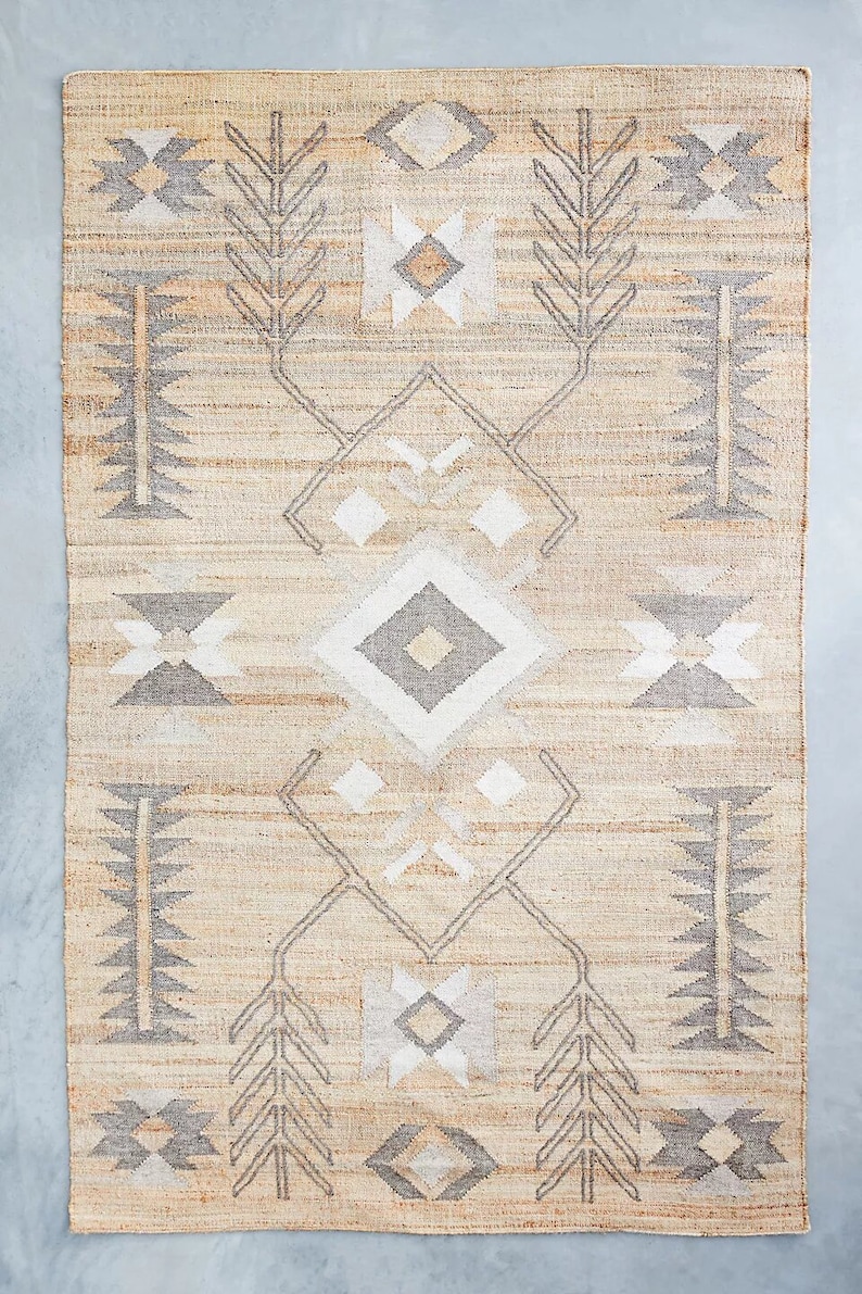 May include: A beige and gray area rug with a geometric pattern. The rug has a diamond shape in the center with smaller diamond shapes on either side. The rug has a fringe border.