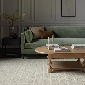 May include: A light green velvet sofa with brown throw pillows sits in a room with grey walls and wood panelling. A wooden coffee table with books and candles sits in front of the sofa. A white rug with a textured pattern covers the floor.