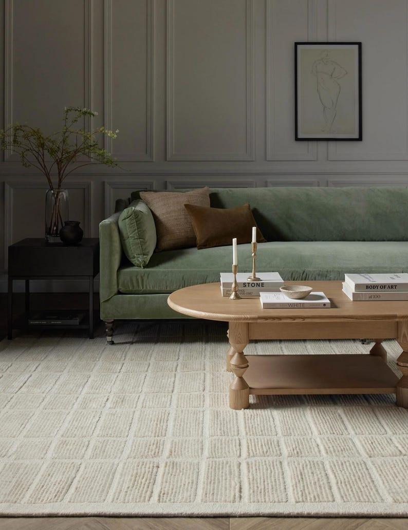 May include: A light green velvet sofa with brown throw pillows sits in a room with grey walls and wood panelling. A wooden coffee table with books and candles sits in front of the sofa. A white rug with a textured pattern covers the floor.