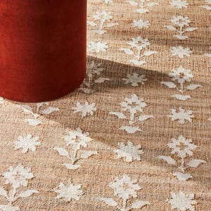 Puede incluir: Una alfombra de yute beige con un patr&oacute;n floral blanco repetido. La alfombra es rectangular y tiene una textura tejida. Una mesa auxiliar cil&iacute;ndrica de color &oacute;xido est&aacute; en primer plano, con una tapa blanca.