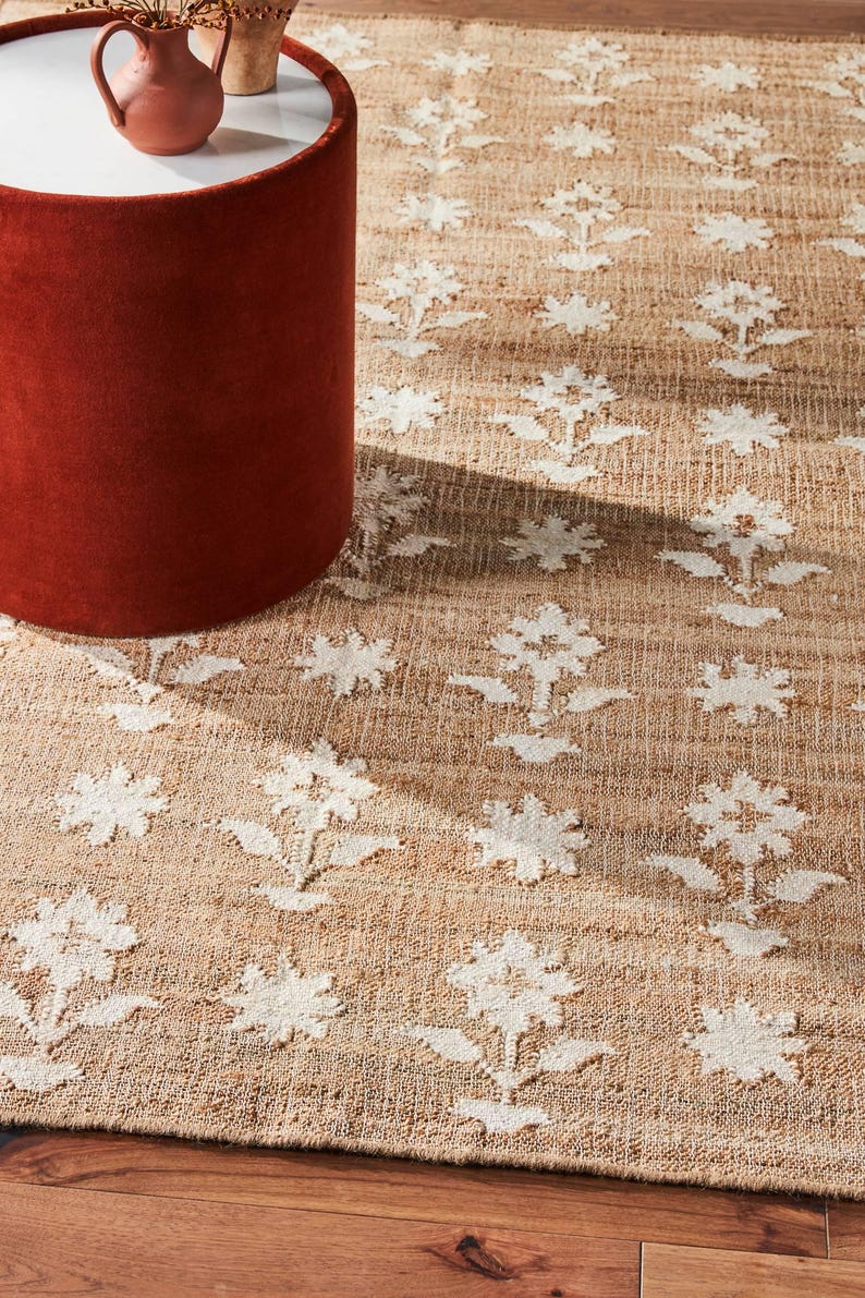 Puede incluir: Una alfombra de yute beige con un patr&oacute;n floral blanco repetido. La alfombra es rectangular y tiene una textura tejida. Una mesa auxiliar cil&iacute;ndrica de color &oacute;xido est&aacute; en primer plano, con una tapa blanca.