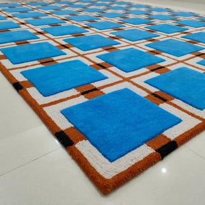 Op de afbeelding: Een groot, rechthoekig vloerkleed met een geometrisch patroon. Het vloerkleed heeft een rasterpatroon van blauwe vierkanten, witte vierkanten, bruine lijnen en zwarte accenten. De rand is warmbruin en de vierkanten zijn gelijkmatig verdeeld.