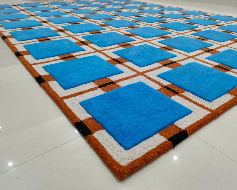 Op de afbeelding: Een groot, rechthoekig vloerkleed met een geometrisch patroon. Het vloerkleed heeft een rasterpatroon van blauwe vierkanten, witte vierkanten, bruine lijnen en zwarte accenten. De rand is warmbruin en de vierkanten zijn gelijkmatig verdeeld.