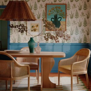 May include: A dining room with a light blue wall and a patterned wallpaper featuring ferns. A wooden table with four chairs sits in the center of the room. The chairs are made of woven wicker and have a light brown finish. A green vase with a floral print sits on the table.