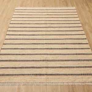 May include: A rectangular area rug featuring a woven texture with alternating horizontal stripes in natural beige and dark charcoal gray. The rug has a flat-weave construction and is finished with short, straight fringe along the top and bottom edges.