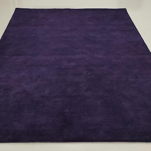 Puede incluir: Una alfombra rectangular de color púrpura oscuro con una textura suave y tupida. La alfombra está colocada sobre un suelo de baldosas de color claro. El color es uniforme en toda la superficie, creando un aspecto sólido y elegante. Mide unos 200 cm x 300 cm.
