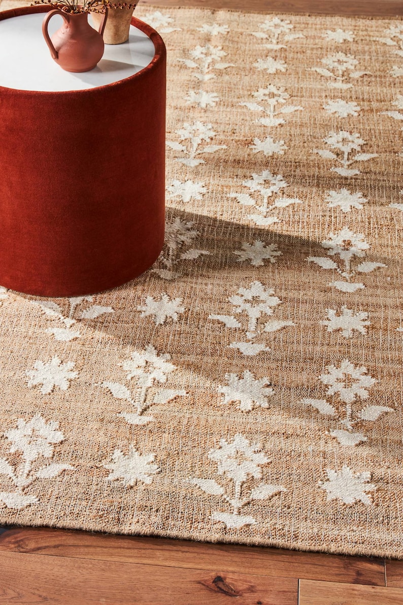 May include: A brown and white patterned jute rug with a floral design. The rug is on a wooden floor and a red cylindrical stool is in the foreground.