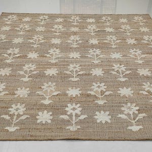 Puede incluir: Una alfombra rectangular de yute beige con un estampado floral repetido en blanco roto. La alfombra est&aacute; hecha de fibras naturales y tiene una textura tejida. El dise&ntilde;o floral presenta flores estilizadas y elementos en forma de estrella.