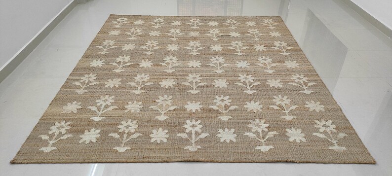 Puede incluir: Una alfombra rectangular de yute beige con un estampado floral repetido en blanco roto. La alfombra est&aacute; hecha de fibras naturales y tiene una textura tejida. El dise&ntilde;o floral presenta flores estilizadas y elementos en forma de estrella.