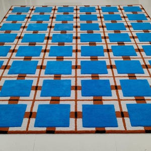Op de afbeelding: Een groot rechthoekig tapijt met een geometrisch patroon. Het tapijt heeft een rasterontwerp met afwisselende vierkanten in helderblauw en bruin, tegen een witte achtergrond. Het tapijt is gemaakt van een zacht, pluche materiaal.