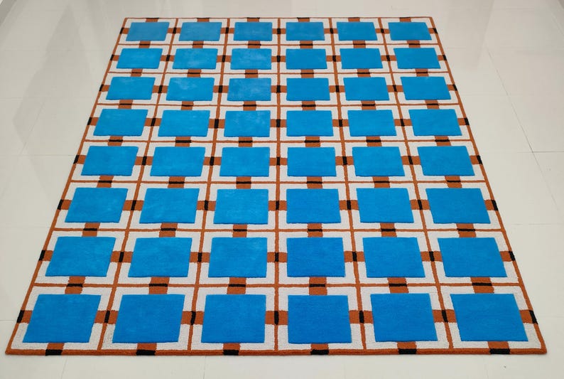 Op de afbeelding: Een rechthoekig tapijt met een geometrisch patroon. Het tapijt heeft een rasterontwerp met afwisselende blauwe vierkanten en bruine en zwarte lijnen op een witte achtergrond. Het tapijt is gemaakt van een zacht materiaal.