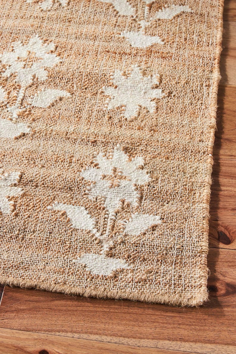 Puede incluir: Una alfombra rectangular de yute con un estampado floral. La alfombra presenta un fondo beige natural con dise&ntilde;os de flores blancas. La alfombra est&aacute; colocada sobre un suelo de madera, mostrando su textura y dise&ntilde;o. Los bordes de la alfombra est&aacute;n cuidadosamente acabados.
