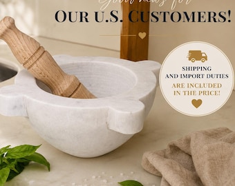 May include: A white marble mortar and pestle set featuring a smooth, rounded bowl with four protruding handles and a light-colored wooden pestle. The mortar has a polished finish and is positioned on a light-colored surface with green basil leaves.