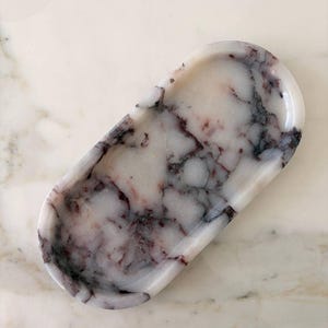 Puede incluir: Una bandeja de forma ovalada hecha de mármol blanco con vetas prominentes de color gris oscuro y burdeos intenso en toda la superficie. La bandeja presenta un acabado liso y pulido con una base poco profunda y bordes elevados, mostrando los patrones naturales de la piedra.