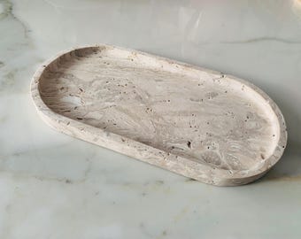 May include: An oval-shaped, beige decorative tray with a marbled pattern. The tray is made of a solid material and has a smooth, slightly textured surface. The tray is designed to hold small items.