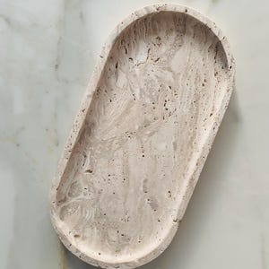 Könnte beinhalten: Eine ovale, beige Steinschale mit strukturierter Oberfläche. Die Schale hat eine geringe Tiefe und abgerundete Kanten. Der Stein hat ein marmoriertes Aussehen mit Farb- und Musterunterschieden. Die Schale ist für dekorative oder funktionale Zwecke konzipiert.