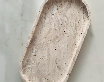May include: An oval-shaped, beige stone tray with a textured surface. The tray has a shallow depth and rounded edges. The stone has a marbled appearance with variations in color and pattern. The tray is designed for decorative or functional use.