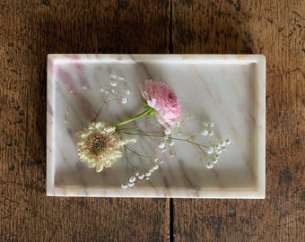 May include: A rectangular white marble tray with subtle pink veining. Resting on the surface of the tray are a single pink ranunculus flower, a pale yellow scabiosa flower, and several small sprigs of white baby's breath flowers.