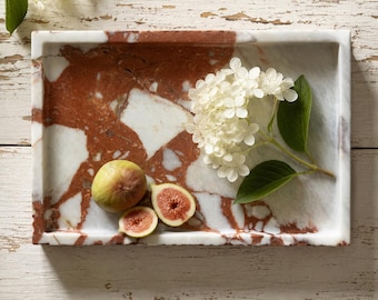 May include: A rectangular marble tray featuring a polished surface with a striking pattern of deep red and white veining. Resting on the tray are a single whole fig, a halved fig showing its pink interior, and a cluster of white hydrangea flowers.