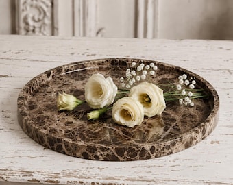 May include: A circular brown marble tray with a polished, veined surface and a raised edge. Three cream-colored eustoma flowers and small sprigs of white baby's breath are arranged on the center of the tray, resting on the smooth stone surface.
