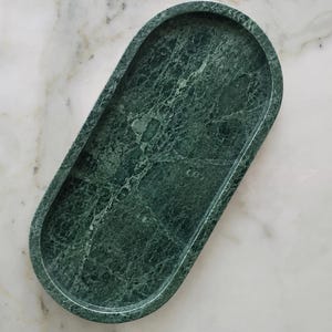 Può includere: Un vassoio di forma ovale realizzato in marmo verde scuro con venature bianche naturali. Il vassoio presenta una superficie liscia e lucida e un bordo rialzato, creando un bacino poco profondo per contenere piccoli oggetti.