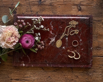 May include: A rectangular dark red marble tray featuring a gold-toned oval pendant necklace, a simple gold band ring, and a pair of gold hoop earrings. A bouquet of a large pale pink carnation and a vibrant magenta ranunculus sits to the left.