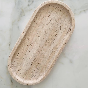 Pode incluir: Bandeja ovalada bege em pedra de travertino. A bandeja tem um design raso e curvo com uma superfície texturizada e uma cor clara e natural. Adequada para pequenos objetos, com cerca de 25 cm de comprimento.