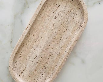 May include: A beige, oval-shaped tray made of travertine stone. The tray has a shallow, curved design with a textured surface and a light, natural color. The tray is suitable for holding small items.